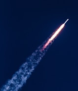 A rocket is ascending into the sky, leaving behind a fiery trail and plume of smoke against a deep blue background.