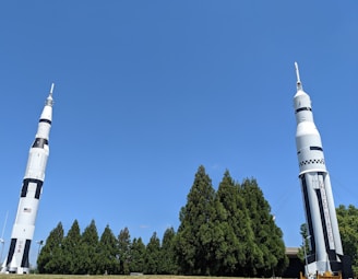 Two large rockets stand upright on a grassy area. The rockets are designed with a white and black color scheme and feature the words 'USA' and 'UNITED STATES' on their sides. They are surrounded by a row of tall green trees, under a clear blue sky.